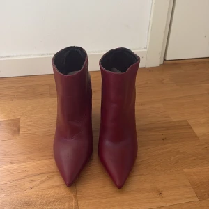 Vinröda stövletter med klack - Snygga vinröda stövletter i skinn med spetsig tå och hög blockklack. Skorna har en stilren design och är perfekta för dig som vill sticka ut med färg. Passar till både jeans och kjol för en trendig look.