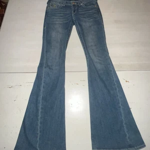 True Religion joey blå bootcut jeans 26 - Snygga blå jeans från True Religion med klassiska kontrastsömmar och ikoniska bakfickor med lock och guldfärgade knappar. Modellen har bootcut-ben och låg midja. Jeansen är i mjukt denimtyg och har en riktigt schysst vintagekänsla.