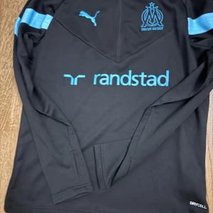 Marseille långärmad träningströja Puma - Snygg svart långärmad fotbollströja från Olympique de Marseille med ljusblå detaljer och logga. Tröjan har halv dragkedja, Drycell-material och tryckta sponsorloggor. Perfekt för träning eller när du vill representera ditt lag.