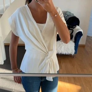 Vit omlottopp med knyt - Snygg vit omlottopp med knyt i midjan och korta ärmar. Toppen har en v-ringad design och ett elegant fall som ger en feminin siluett. Perfekt att styla med jeans eller kjol för en fräsch look.