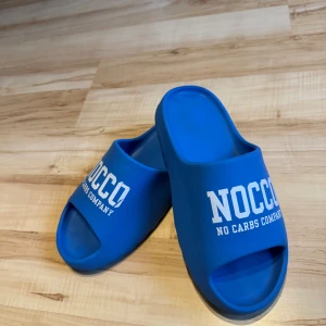 Blåa NOCCO badsandaler - Blåa badsandaler från NOCCO med vit logga framtill. Skorna har öppen tå och platt sula, tillverkade i syntetmaterial. Perfekta att glida runt i på stranden eller vid poolen.