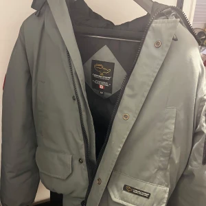 Grå Canada Goose dunjacka med päls - Säljer en grå Canada Goose dunjacka med  pälskant på huvan och klassisk patch på ärmen. Jackan har stora fickor, dragkedja och knappar framtill samt ribbade muddar. Perfekt för kalla dagar och riktigt snygg stil. Jackan har två mindre skador vid kanten av mudden, se bild. Storleken på jackan är XS men passar även S kill storlek, jackan är alltså större i storleken.