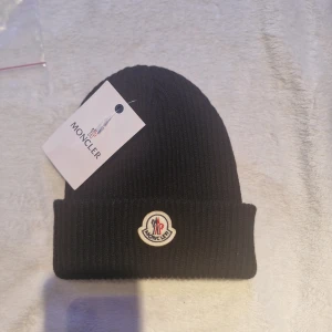 Svart ribbad mössa från Moncler - Svart ribbstickad mössa från Moncler med klassisk uppvikt kant och broderad logotyp framtill. Snygg och enkel design som passar perfekt till kalla dagar. Tillverkad i mjukt material för extra komfort och värme.