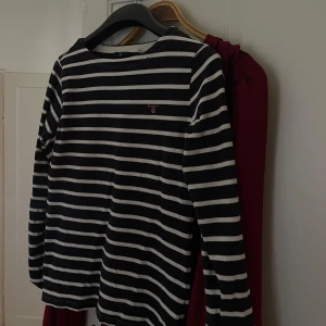 Randig långärmad tröja från GANT - Snygg långärmad tröja från GANT i marinblått och vitt med klassiskt randigt mönster. Tröjan har rund halsringning och broderad GANT-logga i rosa på bröstet. Tillverkad i mjuk bomull, perfekt för en chill och stilren look. Storlek 170 men sitter som en storlek S