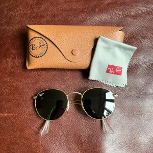 Solglasögon från Ray-Ban - Ett par runda solglasögon från Ray-Ban med mörka linser och tunna, guldfärgade metallbågar (”Ray-Ban Round Metal RB3447”). Använda endast ett fåtal gånger och i mycket fint skick, utan repor eller märken. Brunt hårt fodral och putsduk från Ray-Ban ingår.