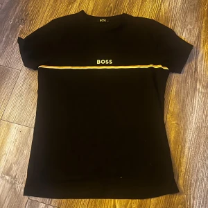 Svart BOSS t-shirt med gul rand - Svart t-shirt från BOSS med korta ärmar och rund hals. Framtill finns en gul och vit vågig rand samt BOSS-logga broderad på bröstet. Skön passform och klassisk stil, perfekt till jeans eller shorts.