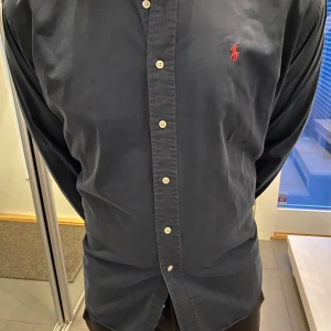 Mörkblå skjorta från Polo Ralph Lauren - Klassisk mörkblå skjorta från Polo Ralph Lauren i 100% bomull. Skjortan har knappar framtill, lång ärm och den ikoniska röda broderade loggan på bröstet. Modellen heter Blaire och har en normal passform som funkar till många olika stilar.