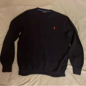 Mörk blå tröja från Polo Ralph Lauren - Klassisk svart tröja från Polo Ralph Lauren med rund hals och den ikoniska broderade loggan i rött på bröstet. Tröjan har lång ärm och är tillverkad i mjuk bomull, perfekt för en clean och stilren look.