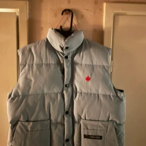 Ljusblå dunväst från Canada Goose - Ljusblå dunväst från Canada Goose med hög krage, stora fickor framtill och broderat rött lönnlöv på bröstet. Västen har tryckknappar och är tillverkad i polyester och bomull med dunfyllning. Perfekt för kalla dagar när du vill hålla stilen.