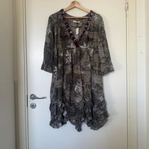 Odd Molly mönstrad kortklänning - Unik kortklänning i grått och beige med vitt blommönster, broderade fjärilar och volangkant. V-ringad med lila sammetsband och stora paljetter. Lätt och flowy bomull, trekvartsärm och avslappnad passform. Från märket Odd Molly i storlek 2 som enligt deras storleksguide motsvarar en storlek M. Mycket fint skick, jag hittar inga synlig anmärkningar eller defekter. 