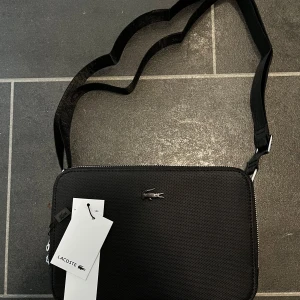 Svart axelväska från Lacoste - Snygg svart axelväska från Lacoste med justerbart band där loggan är invävd. Väskan har silverfärgad dragkedja, diskret Lacoste-krokodil framtill och innerfack med dragkedja. Tillverkad i slitstark syntet med stilren, boxig form.