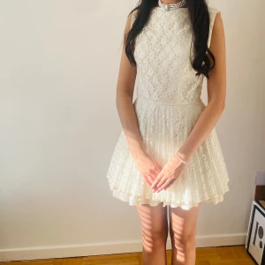 Vit spetsklänning med öppen rygg - Superfin vit kortklänning i spets med blommigt mönster och plisserad kjol. Hög halsringning fram och djup v-ringad öppen rygg med knytband. Perfekt för sommarens speciella tillfällen. Använd endast en gång! Passar xs/small