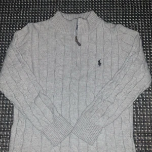 Grå stickad tröja Polo Ralph Lauren - Kabelstickad grå tröja från Polo Ralph Lauren med halv dragkedja och broderad logga på bröstet. Hög krage och långa ärmar, perfekt för lager-på-lager. Klassisk och stilren design som funkar året runt.