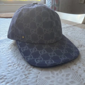 Gucci keps med GG-mönster, blå - Snygg blå keps från Gucci med klassiskt GG-mönster över hela kepsen. Justerbar rem baktill i läder och gulddetalj. Tillverkad i bomullscanvas med ljus insida. Perfekt för dig som vill sticka ut med en lyxig accessoar.
