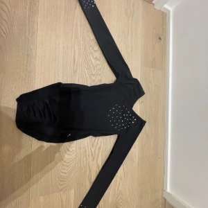 Svart långärmad body men paljetter✨✨ - Svart långärmad tränings body för barn. Med v-ringning och glittriga stenar på ärmarna och  bröstkorgen.stretchigt tyg med resor med till. Perfekt för dans,ballet,gymnastik eller annat….         Står ingen storlek men skulle gissa 134-140/128-134.