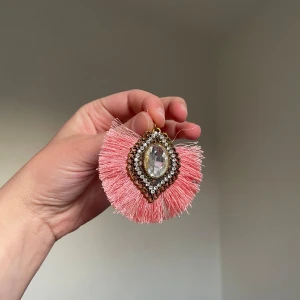 Stort rosa berlock med strass och fransar  - Unikt berlock med rosa fransar och en stor glittrig strassten i mitten, omgiven av små gnistrande stenar. Formen är droppformad och kanten är dekorerad med guldfärgade detaljer. Perfekt statement piece för dig som vill sticka ut.