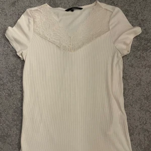 Vit ribbad topp med spetsdetalj - Supersöt vit ribbad topp med korta ärmar och v-ringning dekorerad med spets upptill. Toppens material är mjukt och stretchigt, perfekt för en chill och stilren look. Spetsen ger en extra touch och den sitter snyggt figurnära.