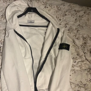 Vit hoodie från Stone Island - Vit hoodie från Stone Island i storlek M med svart dragkedja och klassisk logga på vänster ärm. Hoodien har luva, två fickor framtill och är tillverkad i mjuk bomull. Perfekt för dig som gillar clean och stilren streetwear. Väldigt fet och fresh. Tveka inte att skriva.