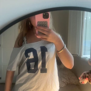 Vit offshoulder t-shirt med tryck - Vit t-shirt med blått tryck av siffran 16 framtill. Modellen är offshoulder och har korta ärmar. T-shirten är loose fit och tillverkad i mjukt bomullstyg, perfekt för en chill och avslappnad stil.