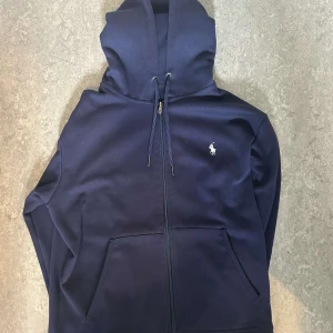 Mörkblå zip hoodie från Ralph Lauren - Mörkblå hoodie från Ralph Lauren med dragkedja och klassisk broderad logga på bröstet. Tröjan har huva med snörning och stora fickor framtill. Perfekt för dig som gillar stilren och enkel design med premiumkänsla.