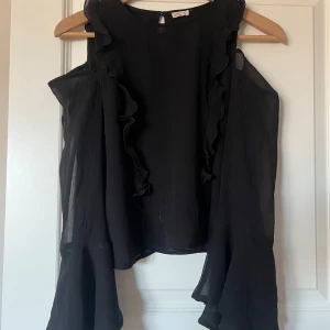 Svart blus från Hollister - Säljer en elegant svart blus från Hollister med volangdetaljer. Blusen är öppen i axlarna och har en luftig design som passar perfekt för varmare dagar. Perfekt för att klä upp eller ner beroende på tillfälle.