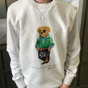 Vit Ralph Lauren tröja med Björn  - Säljer en vit sweatshirt från Ralph Lauren med det ikoniska Polo Bear-motivet på framsidan. Tröjan har långa ärmar och en rund halsringning. Perfekt för en avslappnad och stilren look. 🐻