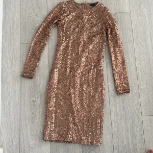 Glittrig klänning med paljetter - Snygg glittrig klänning i guld med paljetter och långärmad design. Klänningen har en dragkedja i ryggen och ett geometriskt mönster som ger en festlig look.