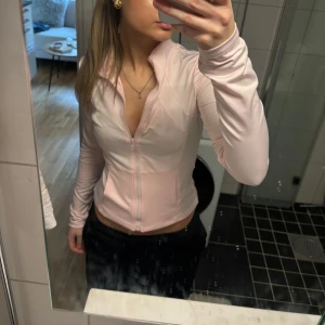 Ljusrosa zip-tröja - Säljer en ljusrosa träningströja med lång ärm och dragkedja framtill. Perfekt för en sportig look eller en avslappnad dag. Tröjan har en figurnära passform och är tillverkad i ett mjukt material.