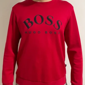Röd tröja från Hugo Boss - Säljer en ÄKTA snygg röd tröja från Hugo Boss med svart logotyp på framsidan. Tröjan har långa ärmar och en klassisk rund halsringning. Tröjan har storlek XL men har passformen för L då det är bomulls material. Tröjan har QR kod som kopplas till Hugo Boss