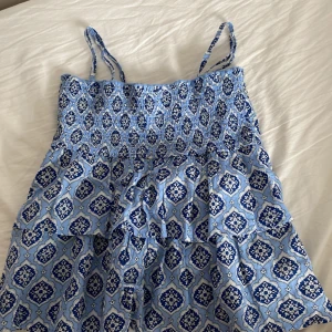 Blå mönstrad topp från Gina Tricot - Säljer en söt blå topp från Gina Tricot med ett detaljerat mönster i vitt och mörkblått. Toppen har smala axelband och volanger. Perfekt för varma sommardagar!