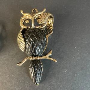 Uggleformat halsband i guld och svart - Unikt halsband med en uggla i guld och svart. Ugglan har ett detaljerat mönster och hänger från en gren. Kedja till smycket saknas 