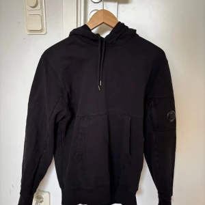 Svart hoodie från C.P. Company - Snygg svart hoodie från C.P. Company. Perfekt för en stilren och bekväm look. Märkeslogga på ärmen och text på luvkanten. Inga defekter. Storleken är M men den är lite liten i storleken. Nypris 2000kr.