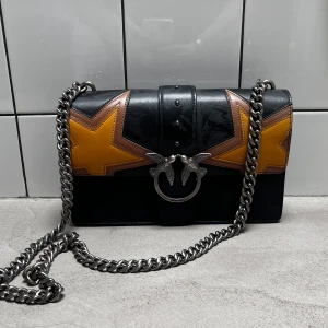 Svart axelväska med stjärndetaljer - Snygg svart axelväska från Pinko med kedjerem och coola gula stjärndetaljer. Väskan har en unik spänne framtill och är perfekt för att ge din outfit en edgy touch. Lite dammig😄