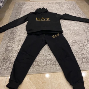 Svart hoodie från Emporio Armani - Snygg svart hoodie från Emporio Armani med guldtryck på bröstet. Tröjan har en klassisk design med huva och en stor ficka framtill. Perfekt för en stilren och bekväm look.