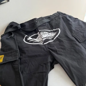 Cortize - Säljer ett par svarta cargopants med coolt tryck på framsidan. Byxorna har flera praktiska fickor och en justerbar midja med bälte. Perfekta för en avslappnad stil.