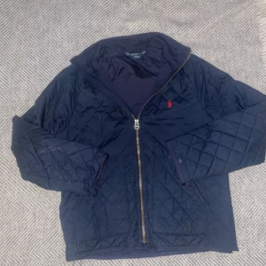Mörkblå quiltad jacka från Polo Ralph Lauren - Säljer en snygg mörkblå quiltad jacka från Polo Ralph Lauren med dragkedja och broderad logga på bröstet. Jackan har långa ärmar och är perfekt för kyligare dagar. Passar bra till både vardag och mer uppklädda tillfällen.