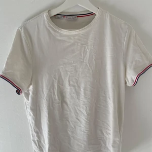 Vit t-shirt från Moncler - Säljer en stilren vit t-shirt från Moncler med slim fit passform. T-shirten har korta ärmar med röd och blå randig detalj vid ärmsluten. Perfekt för en avslappnad och snygg look.