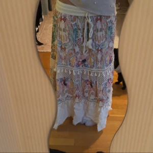 Färgglad långkjol med spetsdetaljer - Säljer en vacker långkjol med färgglatt blommönster och spetsdetaljer. Kjolen har en bekväm elastisk midja med knytband och tofsar. Perfekt för en bohemisk look!