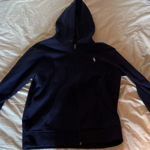 Mörkblå hoodie från Polo Ralph Lauren - Säljer en stilren mörkblå hoodie från Polo Ralph Lauren med dragkedja och klassisk logga på bröstet. Perfekt för en avslappnad look. Den har en justerbar huva och är tillverkad i mjukt material för extra komfort. Perfekt inför sommaren om man vill vara stilig