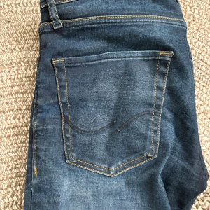Blå jeans från Jack & Jones - Snygga blå jeans från Jack & Jones med klassisk femficksdesign och kontrastsömmar. De har en knappgylf och en läderpatch med logotyp baktill. Perfekta för en avslappnad stil.