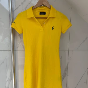 Gul klänning från Ralph Lauren - Snygg gul klänning från Ralph Lauren med klassisk pikédesign och blå logga på bröstet. Perfekt för en somrig look med krage och korta ärmar. Märkesnamnet är broderat på ärmen.