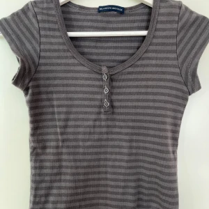 Grå randig topp från Brandy Melville - Säljer en grå randig topp från Brandy Melville. Toppen har korta ärmar och tre knappar framtill. Perfekt för en avslappnad stil.
