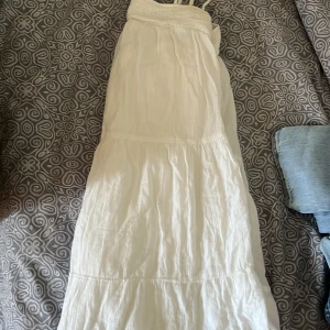 Vit långklänning med smockdetaljer - Säljer en  vit kortklänning  med smala axelband och smockdetaljer upptill. Klänningen har en luftig och flödande design, perfekt för varma dagar. Materialet är lätt och bekvämt.