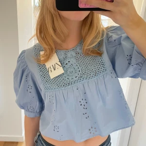 Blå blus med spetsdetaljer - Säljer en söt blå blus från Zara med spetsdetaljer på bröstet och broderade hålmönster. Blusen har puffiga kortärmar och en croppad passform. Perfekt för en somrig look!