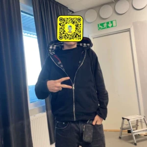 Svart hoodie med dragkedja - Säljer en svart hoodie med dragkedja och rutigt foder i luvan. Perfekt för en avslappnad stil. Den har långa ärmar och en bekväm passform.
