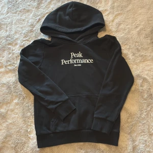 Svart hoodie från Peak Performance - Säljer en svart hoodie från Peak Performance med vit logga på bröstet. Tröjan har en klassisk känguruficka och ribbade muddar vid ärmslut och nederkant. Perfekt för en avslappnad stil.
