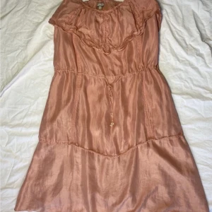 Rosa klänning från Noa Noa - Säljer en söt rosa klänning från Noa Noa med volangdetaljer och knytband i midjan. Klänningen har en mjuk och glansig finish som ger en elegant touch. Perfekt för somriga dagar eller speciella tillfällen.