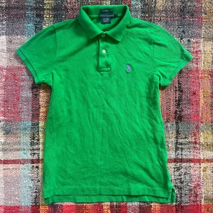 Grön pikétröja från Ralph Lauren - Säljer en snygg grön pikétröja från Ralph Lauren med klassisk krage och knappar framtill. Tröjan har en broderad logotyp på bröstet i lila. Perfekt för en stilren look!
