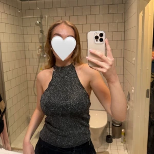 Glittrig ärmlös topp - Snygg glittrig ärmlös topp med knytning i nacken. Perfekt för en kväll ute eller festliga tillfällen. Toppen har en tight passform och är i en mörk färg som ger en elegant look.