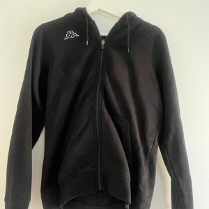 Svart hoodie från Kappa - Säljer en klassisk svart hoodie från Kappa med dragkedja och huva. Tröjan har långa ärmar och en liten logga på bröstet. 